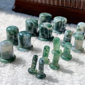 May include: A collection of moss agate plugs in various sizes, each featuring a smooth, cylindrical shape with a flared base. The stones display translucent white and deep green patterns, with unique inclusions resembling moss or foliage throughout.