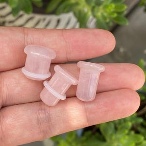 May include: Three pale pink rose quartz ear plugs with a smooth, polished finish. Each piece features a cylindrical body with a flared rim at one end and a small, translucent white rubber O-ring positioned around the shaft to secure the jewelry.