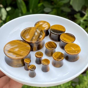 Könnte beinhalten: Eine Sammlung von polierten Ohr-Plugs aus Tigerauge-Stein in verschiedenen Größen, mit einer glatten, konkaven Sanduhrform, ausgeprägten goldbraunen chatoyierenden Bändern und einer glänzenden Oberfläche, angeordnet auf einem weißen Tablett.