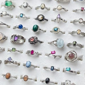 May include: A collection of assorted rings featuring various gemstones and metal bands arranged in rows. The rings include diverse shapes, colors, and settings, such as oval, round, and pear-cut stones in silver-toned metal, displayed on a white surface.