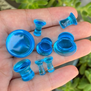 May include: A collection of seven translucent bright blue glass ear plugs of varying sizes. Each piece features a flared design with a concave center, showcasing a smooth, polished finish and a vibrant, uniform cyan color throughout the set.
