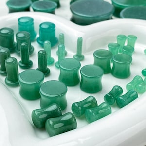May include: A collection of green stone ear plugs and tunnels in various sizes and shapes, including flared and straight designs, arranged on a white heart-shaped tray. The pieces feature smooth, polished surfaces with varying shades of green.