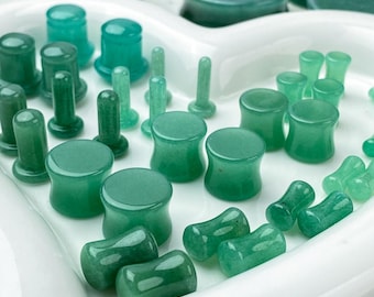 May include: A collection of green stone ear plugs and tunnels in various sizes and shapes, including flared and straight designs, arranged on a white heart-shaped tray. The pieces feature smooth, polished surfaces with varying shades of green.