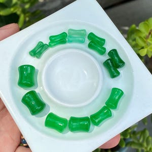 May include: A collection of twelve small, smooth, green, hourglass-shaped ear plugs arranged in a circle on a white, square-shaped tray. The plugs vary in shade from light mint green to a deep, vibrant emerald green, each featuring a concave center.