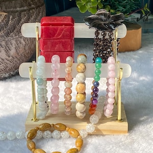 Puede incluir: Un expositor de joyas de madera con varias pulseras de cuentas. Las pulseras presentan una gama de colores, incluyendo rojo, rosa, verde y marrón. También se ve una flor negra tallada y una pulsera con cuentas de madera.