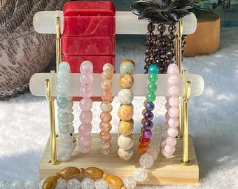Op de afbeelding: Een houten sieradenstandaard met diverse armbanden met kralen. De armbanden hebben een reeks kleuren, waaronder rood, roze, groen en bruin. Een gesneden zwarte bloem en een armband met houten kralen zijn ook zichtbaar.