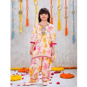 May include: A two-piece outfit consisting of a tunic and matching pants in a light cream fabric with a vibrant yellow and pink floral print. The tunic features a round neckline with colorful embroidery, three-quarter sleeves, and pink tassels.