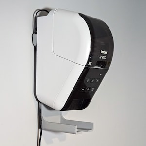 May include: A white and black Brother label printer mounted on a wall. The printer has a curved design with a black top and control panel. A gray bracket supports the printer, and a black cable runs along the wall.