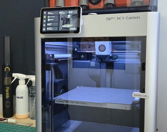 May include: A silver Bambu Lab X1-Carbon 3D printer with a clear front panel, displaying a print bed and internal components. Above, an orange and gray AMS unit holds filament. A touchscreen shows the printer's interface. Additional cleaning supplies are visible.