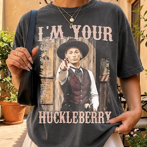 May include: A charcoal gray short-sleeve t-shirt featuring a graphic print of a person in a western-style vest and hat with the text I'M YOUR HUCKLEBERRY. The shirt is paired with a white pleated skirt and a black shoulder bag.