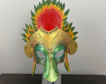 Puede incluir: Una máscara de carnaval verde, dorada y roja con un tocado de plumas. La máscara tiene un diseño detallado con un borde dorado y una cara verde con un hocico largo.