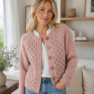 Peut inclure: Un gilet en tricot rose poudré avec un motif torsadé texturé, une encolure ronde et cinq boutons assortis sur le devant. Ce vêtement à manches longues est porté sur une chemise blanche à col et un jean bleu, affichant un design en maille.