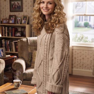 May include: An oversized beige cable-knit sweater featuring a thick, textured pattern with vertical braided cables and ribbed cuffs. The garment has a high neckline and a loose, relaxed fit, constructed from a chunky yarn with a neutral tone.
