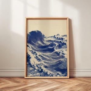 May include: A framed art print featuring a detailed illustration of ocean waves in shades of blue. The artwork is in a wooden frame and leans against a white wall on a wooden floor. The waves are depicted with intricate lines, creating a sense of movement.