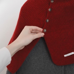 May include: A red knitted cape with a button closure. The cape is a vibrant red color and features several brown buttons down the front. The cape is layered over a gray garment. A hand is shown buttoning the cape.