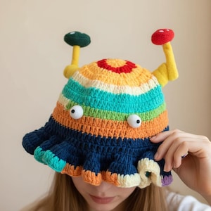 Può includere: Un cappello all'uncinetto colorato e fatto a mano con un design stravagante. Il cappello presenta un motivo a righe nei colori dell'arcobaleno, con due abbellimenti a forma di fungo e occhi mobili. L'orlo è a balze e blu navy.