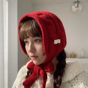 May include: A vibrant red knitted bonnet with a cable knit pattern. The bonnet features a large bow at the neck and two small white decorative accents. The ear flaps are designed to keep the ears warm. A cozy winter accessory.