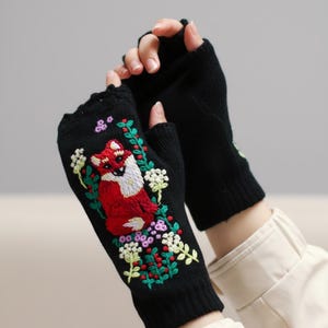 May include: Black fingerless gloves with embroidered fox and floral design. The fox is red and white, surrounded by green leaves, white flowers, and purple berries. The gloves are made of a knitted material.