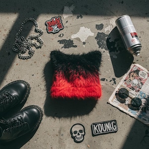 Puede incluir: Un gorro de piel sintética degradado negro y rojo se encuentra sobre una superficie de hormigón, junto a botas negras, una cadena plateada, parches, un periódico y una lata de pintura en aerosol. Los parches incluyen una calavera y el texto "KOUNK".