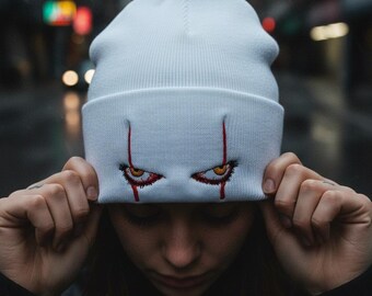 May include: White beanie with an embroidered design of red and yellow eyes with red tear streaks. The beanie is pulled down over the forehead. The background is a blurred city street.