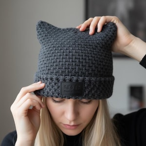 Könnte beinhalten: Eine dunkelgraue Strickmütze mit Katzenohren. Die Mütze hat einen umgeschlagenen Rand und ein schwarzes rechteckiges Etikett mit weißem Text. Die Mütze ist aus einem dicken, strukturierten Garn gefertigt.
