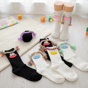 Puede incluir: Calcetines novedosos en blanco y negro, con caras de dibujos animados y cabello trenzado colorido. Un par está enrollado y dos pares están en maniquíes. Los calcetines tienen una textura acanalada.