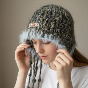 May include: A hand-knitted hat with a textured, speckled gray and green design. The hat features a light blue, fluffy trim and dangling strands with decorative knots. A small label is sewn on the front.
