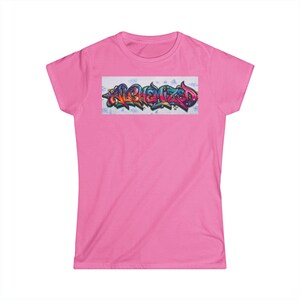 May include: A bright pink short-sleeved t-shirt featuring a rectangular graphic on the chest. The graphic displays the word ALCHEMIZED written in a multi-colored graffiti-style font with shades of blue, purple, orange, and pink against a light background.