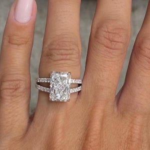 Op de afbeelding: Een rechthoekige diamanten verlovingsring met een gespleten band, bezet met kleinere diamanten. De ring zit aan een hand met lange, lichtroze nagels. De achtergrond is een zachte, neutrale kleur.