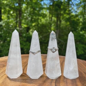 以下が含まれることがあります： 研磨された円錐形をした、透明な水晶のリングホルダー4つ。2つのシルバーリングがホルダーに展示されています。背景は緑の木々のぼやけた景色です。