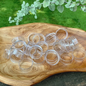 May include: A collection of clear, plastic ring-shaped objects, possibly napkin rings, are arranged in a wooden, organic-shaped bowl. The bowl has a light brown color with a visible wood grain. Green foliage is visible in the background.