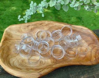 May include: A selection of clear, plastic ring-shaped objects, possibly napkin rings, are arranged in a wooden, organic-shaped bowl. The bowl is light brown with a visible wood grain. Green foliage is visible in the background.