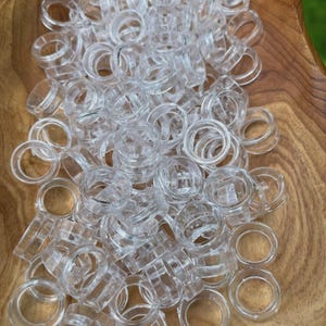 May include: A collection of clear, transparent plastic rings, possibly curtain rings, are piled in a wooden bowl. The rings have a circular design with a slightly textured surface. The wooden bowl has a natural wood grain pattern.