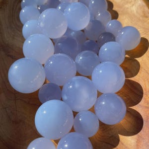 May include: A collection of pale blue, spherical gemstones, possibly chalcedony, are displayed in a wooden bowl. The spheres vary in size and have a translucent quality, reflecting light. The wooden bowl has a warm, natural wood grain.