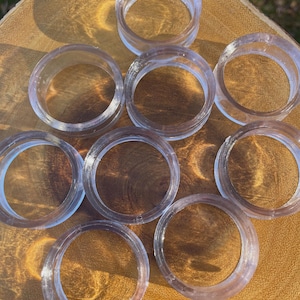 May include: A collection of clear, circular napkin rings. The rings are made of a translucent material and are arranged on a wooden surface. The rings are evenly spaced and appear to be new.