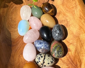May include: A collection of polished gemstone eggs in various colors, including pink, green, blue, and brown, arranged in a wooden bowl. The eggs vary in texture and pattern, with some appearing translucent and others opaque. The arrangement is lit by natural light.