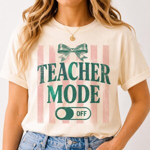 May include: Cream-colored t-shirt with the text "TEACHER MODE OFF" in green, featuring a green bow and pink vertical stripes. The shirt has short sleeves and a relaxed fit.