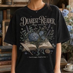 May include: A black short-sleeve t-shirt featuring a graphic print of an open book with a bouquet of blue flowers and a quill pen. The text reads Dearest Reader a literary romance and Lost in pages, found in love with small star accents.