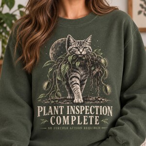 May include: A dark olive green crewneck sweatshirt featuring a graphic of a tabby cat carrying a plant with Venus flytraps. The text reads: PLANT INSPECTION COMPLETE NO FURTHER ACTION REQUIRED. The design includes a moon and starry background.