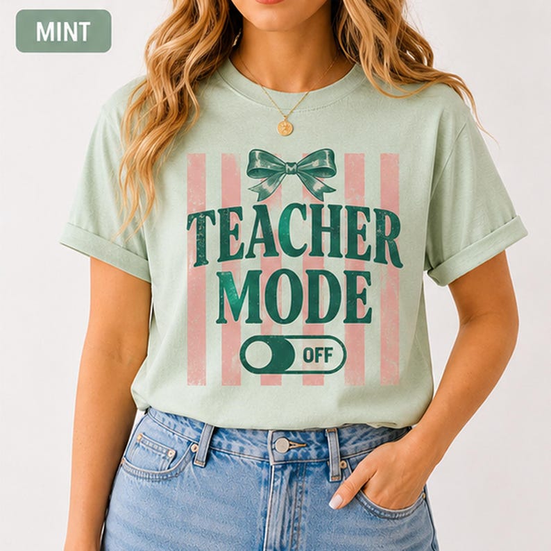 May include: Mint green t-shirt with the text "TEACHER MODE OFF" in dark green. The design includes a bow and pink vertical stripes. The shirt is worn with blue jeans.