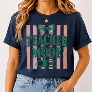 May include: Navy blue t-shirt with a distressed pink and white striped design. The shirt features the text "TEACHER MODE" in green, with a green bow and a switch graphic that says "OFF".