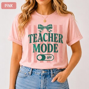 May include: Pink t-shirt with the text "TEACHER MODE OFF" in green, accented by a green bow and vertical stripes. The shirt is a light pink color and has short sleeves. The word "PINK" is in a pink box in the upper left corner.
