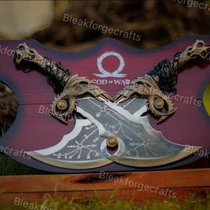 May include: Two crossed, ornate blades with gold accents and intricate designs, mounted on a burgundy wooden plaque. The plaque features the text "GOD OF WAR" and a symbol. The handles are detailed with dragon-like features.