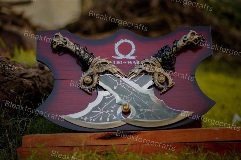 May include: Two crossed, ornate blades with gold accents and intricate designs, mounted on a burgundy wooden plaque. The plaque features the text "GOD OF WAR" and a symbol. The handles are detailed with dragon-like features.