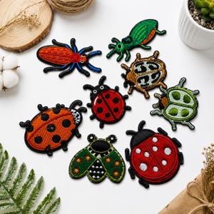 May include: A collection of eight embroidered insect patches featuring various designs, including several red and black ladybugs, a green beetle, a brown patterned beetle, and a green moth-like insect, all with detailed stitching and black borders.