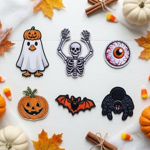 May include: A set of six embroidered patches featuring a ghost wearing a pumpkin hat, a skeleton with raised arms, a bloodshot eyeball, a jack-o'-lantern, a bat with orange wings, and a black spider, all arranged on a white wooden surface.