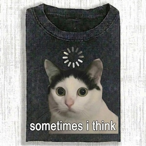 Puede incluir: Camiseta gris oscuro con un gato blanco y negro de ojos grandes y un símbolo de carga. El texto "sometimes i think" está debajo. La camiseta está doblada.