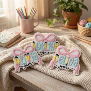 May include: Three custom embroidered patches featuring a pink bow, light blue lettering, and yellow pencil accents. The patches display the names Olivia Jame, Adeline Grace, and Celina Jones in a script font, resting on a cream-colored knit fabric.