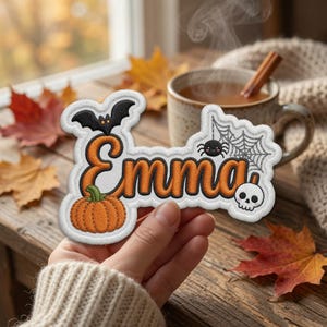 May include: An embroidered patch featuring the name Emma in orange script with a black outline. The design includes a black bat, a small orange pumpkin, a spider with a web, and a white skull, all set against a white background border.