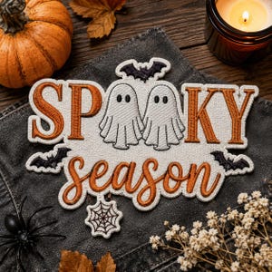 May include: An embroidered patch featuring the text SPOOKY season in orange and white thread. The design includes two white ghosts, three black bats, and a white spiderweb. The patch is placed on a dark gray fabric surface with a pumpkin nearby.